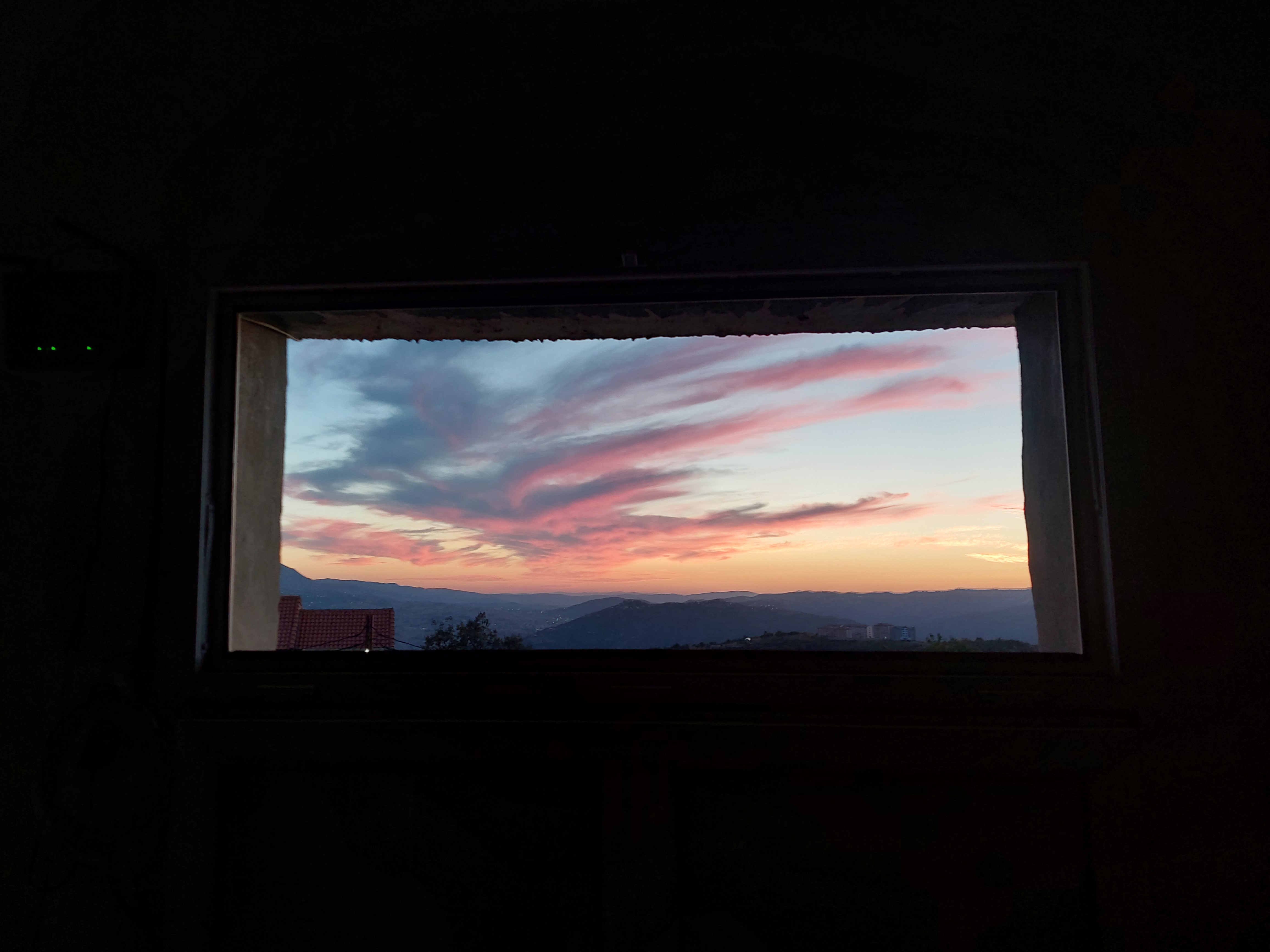Un ciel rose au-dessus des collines, vu depuis une fenêtre dans une pièce sombre.